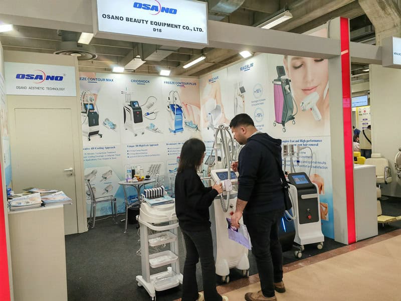 Two people converse at Osano Beauty Equipment's booth, displaying beauty tools and info posters at the 2024 Cosmoprof in Bologna.