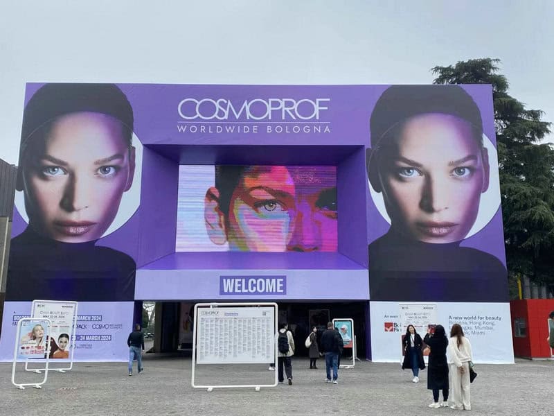 Entrance of Cosmoprof Bologna 2024 with a banner of a woman's face and people walking under a "Welcome" sign.