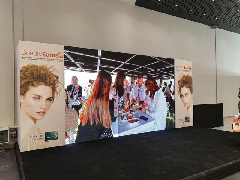 A BeautyEurasia 2024 display features a screen with people chatting at a booth and ads running on panels showcasing the event.