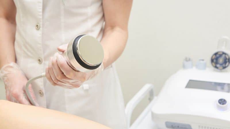 Eine Person im Laborkittel verwendet ein medizinisches Gerät an einem Patienten. Im Hintergrund sind medizinische Geräte zu sehen.