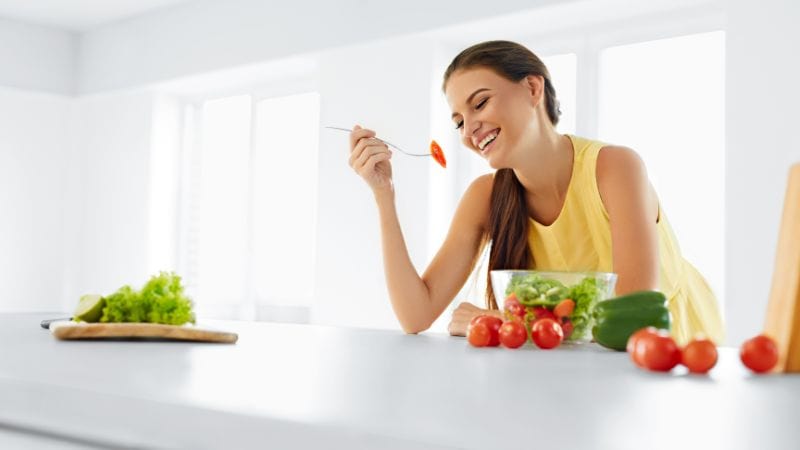 In einer hellen Küche genießt eine Frau im gelben Hemd Salat und frisches Gemüse und ist positiv mit ihrem Körper konfrontiert.