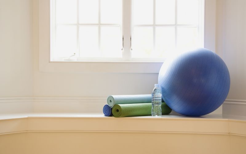 Relaxing home exercise space with stability ball, yoga mats, and water bottle by the window for comfort and wellness.