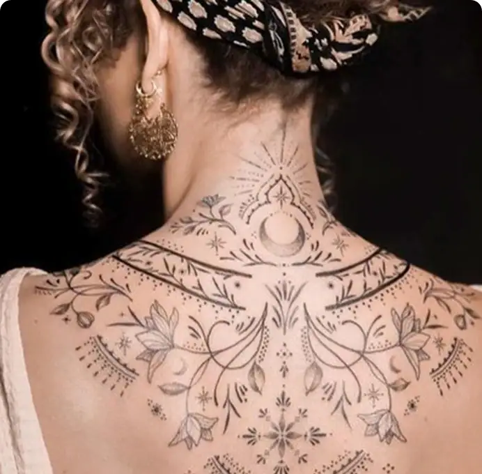 Detailed black ink tattoo on woman's back and neck featuring floral, celestial, and geometric patterns.