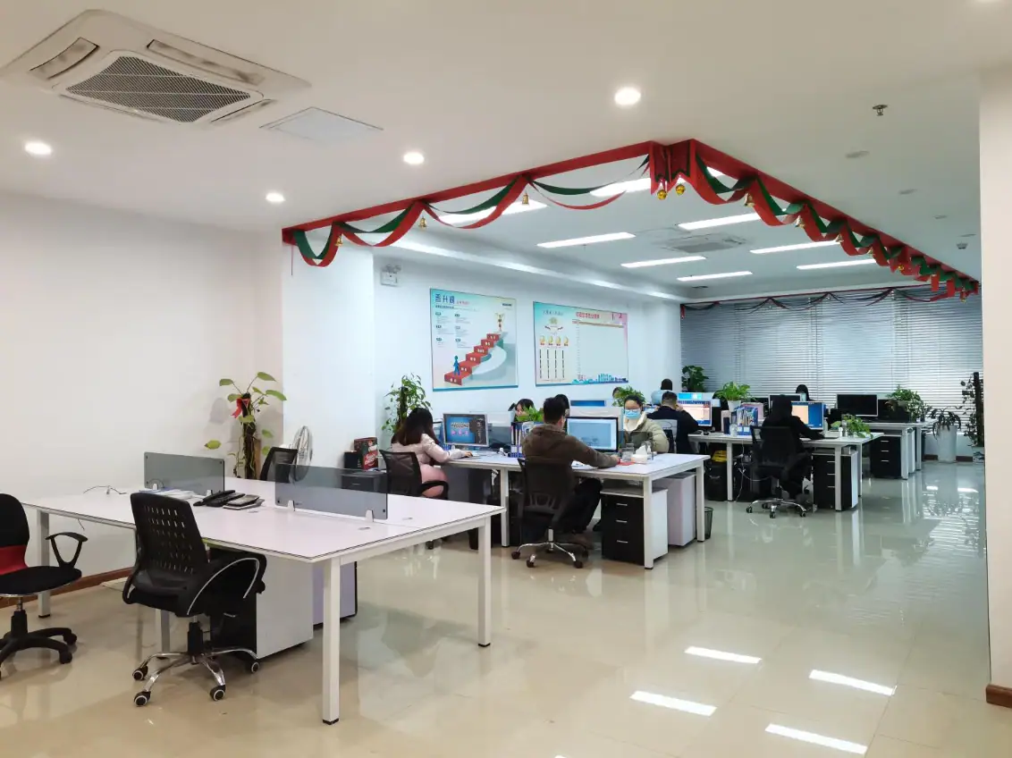 Open-plan office with employees working at desks, decorated with red and green Christmas garlands, bright lighting, and modern furniture.